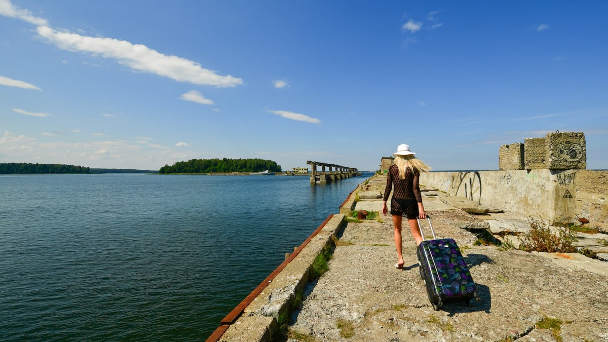 A woman in a black dress and white hat walks on an abandoned pier in Hara, Estonia, under a sunny blue sky.
